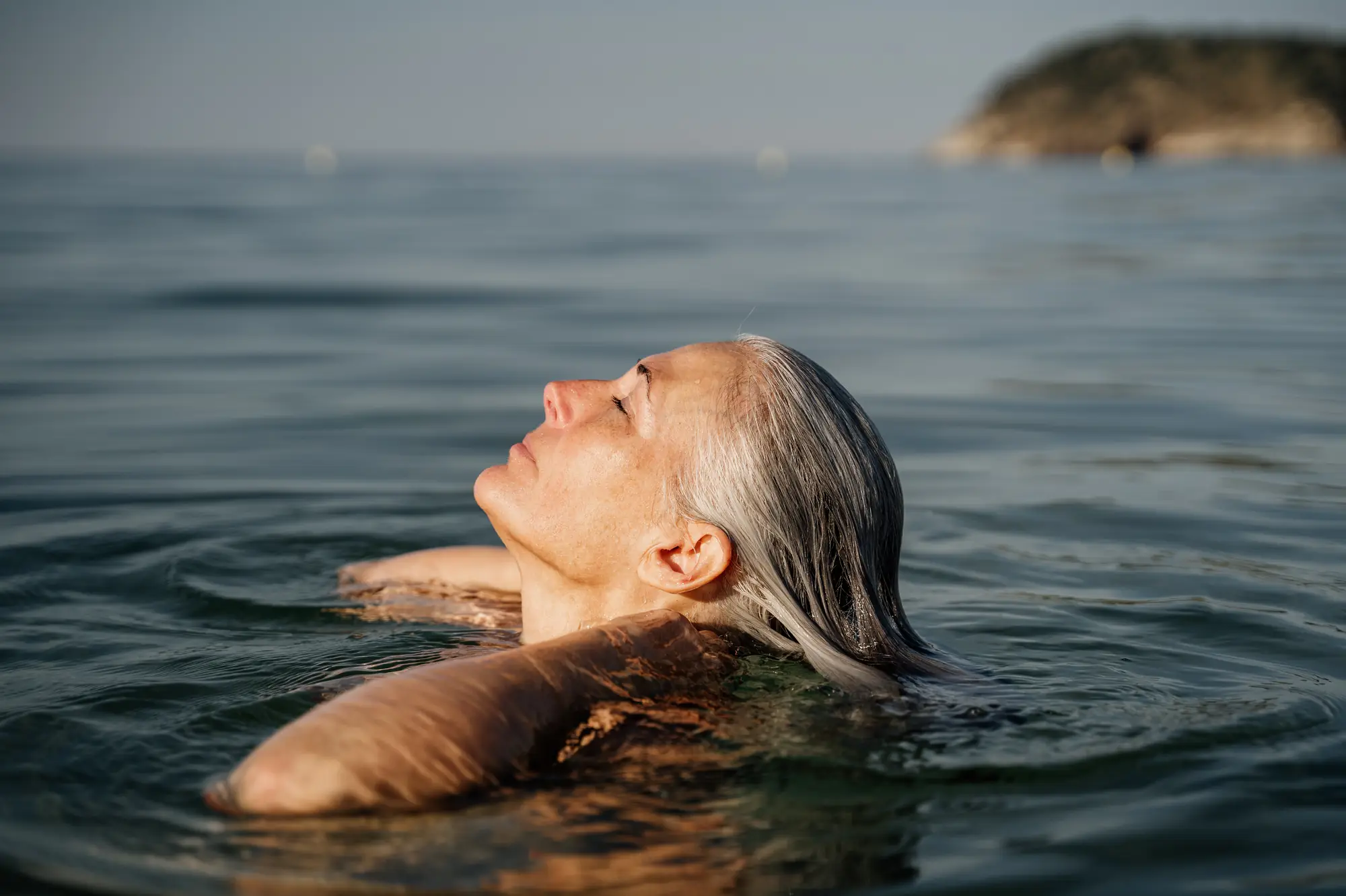A woman floating on her back in calm open water, eyes closed.